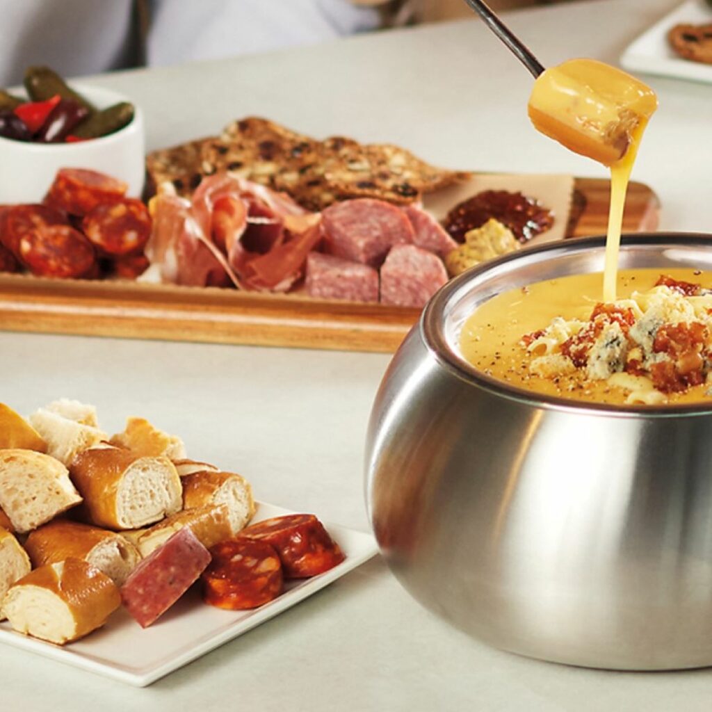 A table with cheese fondue and a selection of charcuterie meats and cheeses and pretzels
